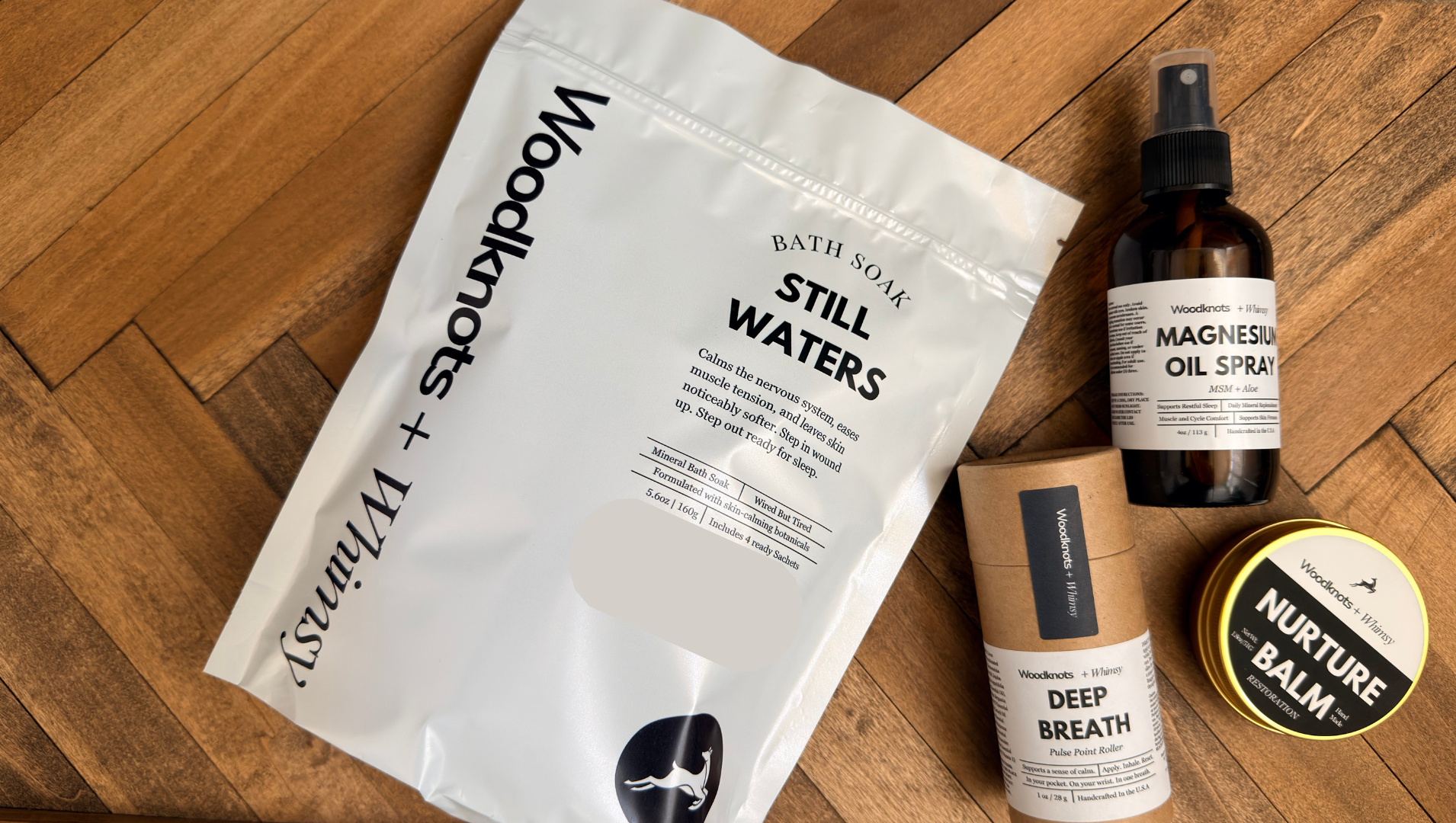 A flat-lay of Woodknots + Whimsy bath and body products on a wooden floor, including a large white 'Still Waters' bath soak pouch, a dark amber magnesium oil spray bottle, a brown paper tube labeled 'Deep Breath', and a round tin labeled 'Nurture Balm'.